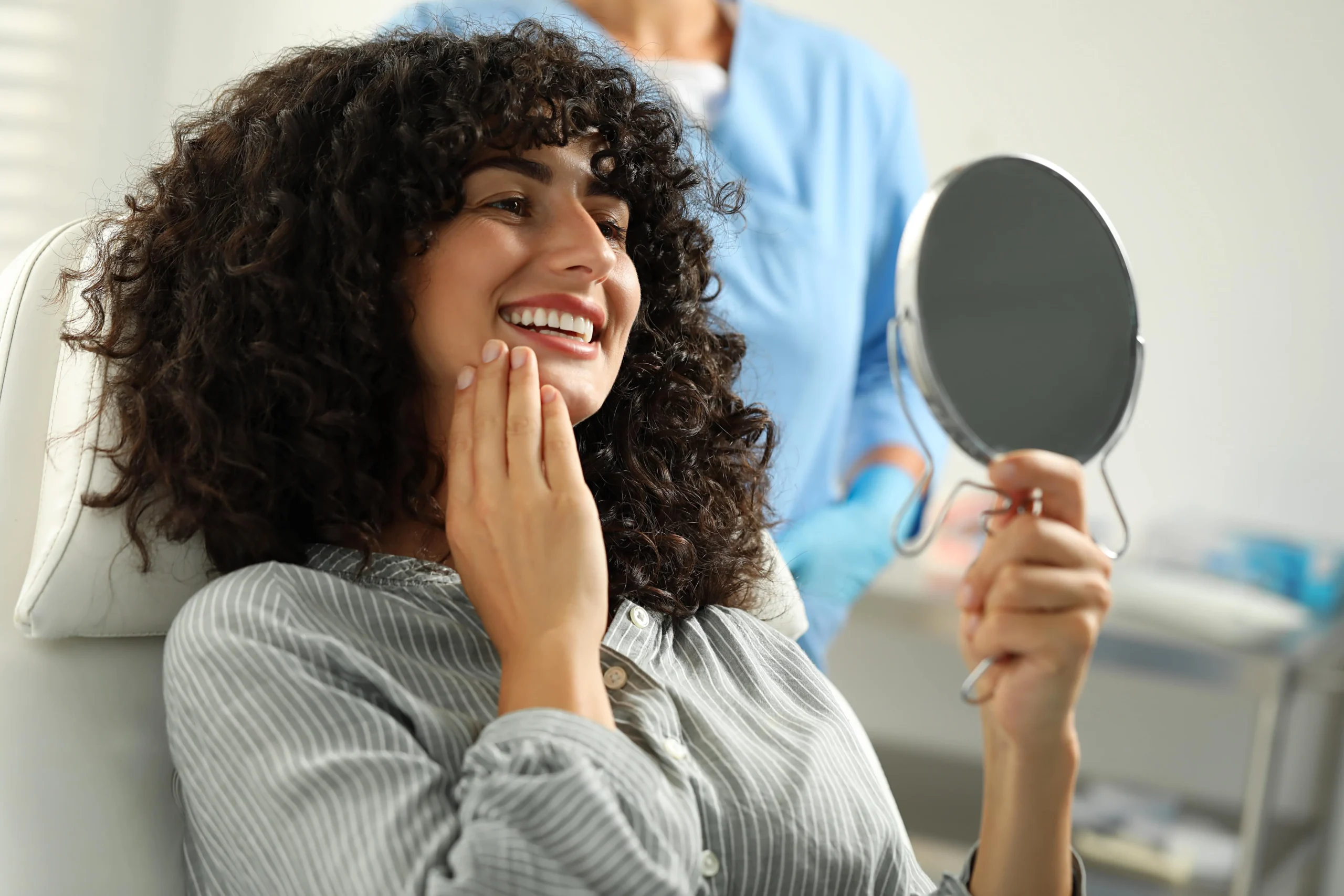 A woman in a dental chair holding a hand held mirror and smiling while looking at her teeth