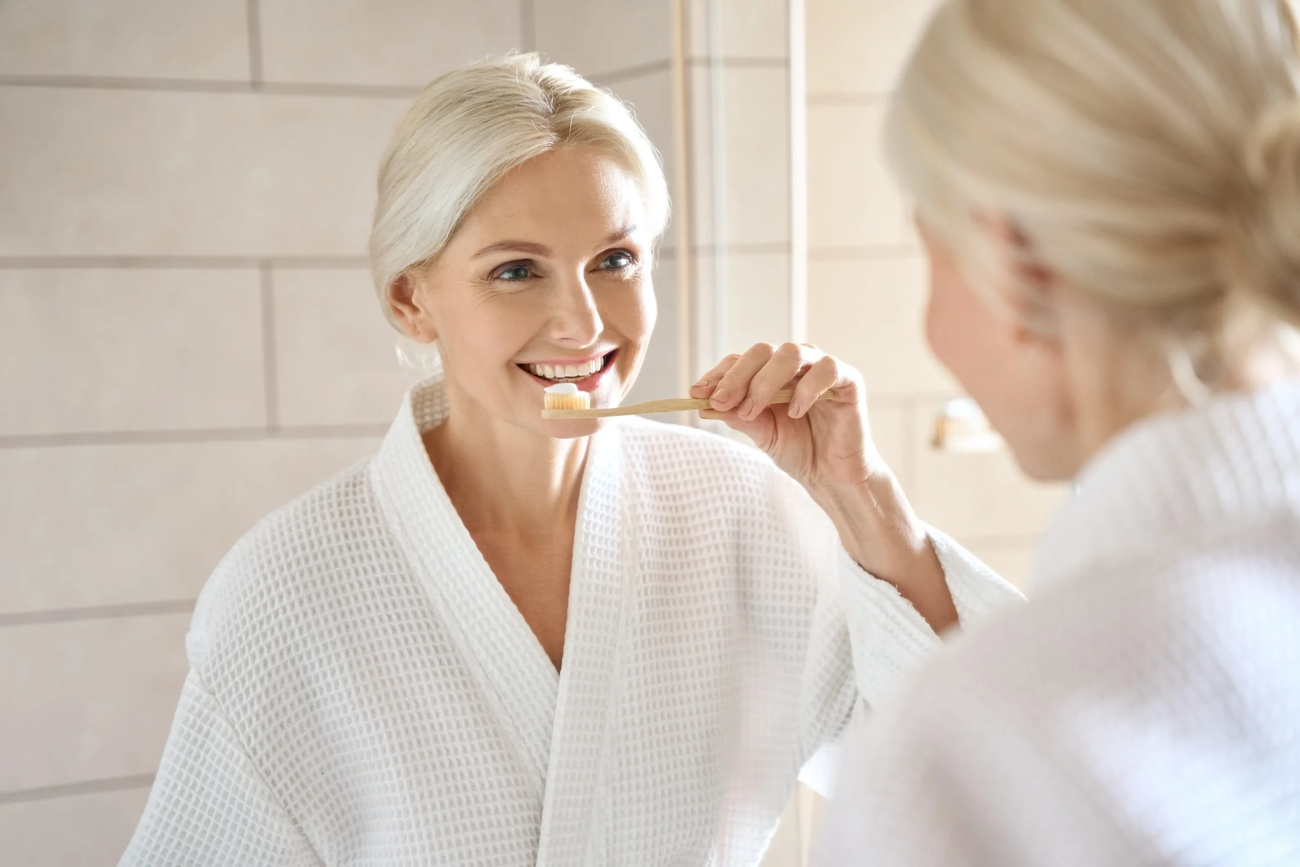 Older woman staring at the mirror brushing her teeth while smiling.